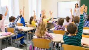 barn sitter vid sina bänkar i ett klassrum och räcker upp handen.